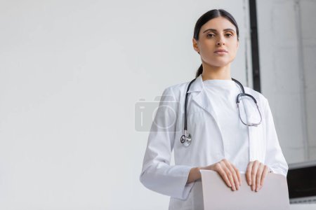 Photo for Low angle view of doctor in white coat holding paper folder and looking at camera in clinic - Royalty Free Image