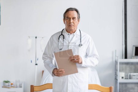 Téléchargez les photos : Senior doctor holding paper folder and looking at camera in clinic - en image libre de droit