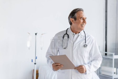 Elderly doctor holding paper folder and smiling in hospital ward 