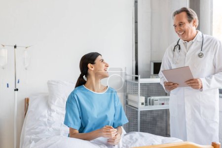 Lächelnder Oberarzt mit Papiermappe in der Nähe von Patient auf Krankenhausstation 