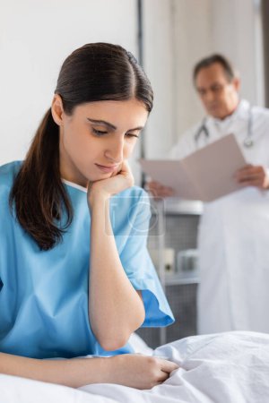 upset patient sitting on bed near blurred doctor in hospital 