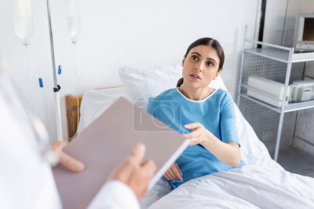 brunette patient talking to blurred doctor with paper folder in hospital ward 