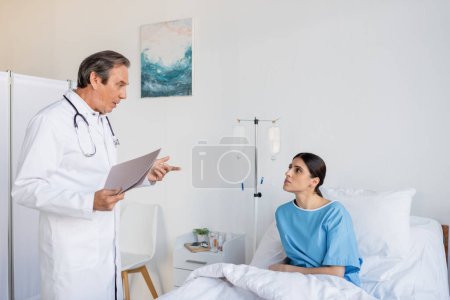 Foto de Senior doctor with paper folder talking to brunette patient in hospital ward - Imagen libre de derechos