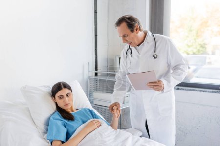Oberarzt mit Papiermappe hält Hand einer brünetten Patientin auf Krankenhausstation 