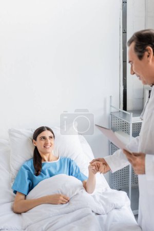 Foto de Smiling patient holding hand of elderly doctor with paper folder in clinic - Imagen libre de derechos