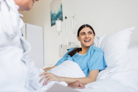 Cheerful patient in gown lying on bed near blurred doctor in clinic 