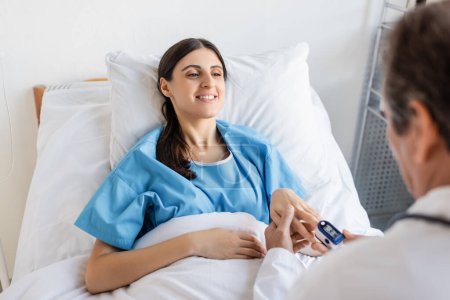 Téléchargez les photos : Smiling patient with pulse oximeter on hand looking at blurred doctor in clinic - en image libre de droit