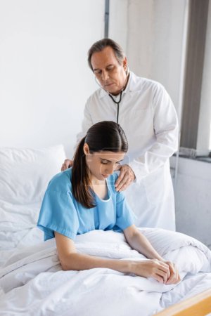 Elderly doctor with stethoscope examining back of patient on bed in clinic  