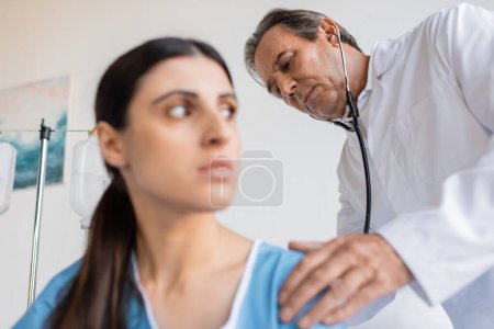 Photo for Senior doctor with stethoscope checking back of blurred patient in clinic - Royalty Free Image