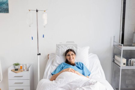 Cheerful patient looking at camera near intravenous therapy in clinic 