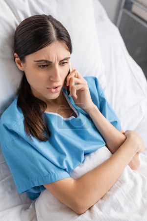 High angle view of worried patient talking on smartphone on bed in hospital 