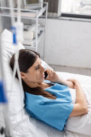 Patient spricht auf Smartphone, während er im Bett liegt, in der Nähe einer verschwommenen intravenösen Therapie im Krankenhaus 
