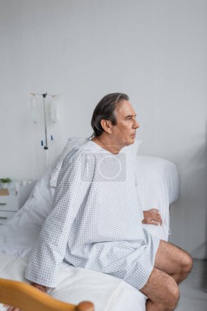 Photo for Grey haired patient in gown sitting on bed in hospital - Royalty Free Image