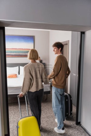 Foto de Young man looking at blonde girlfriend while standing with travel bags in hotel room - Imagen libre de derechos