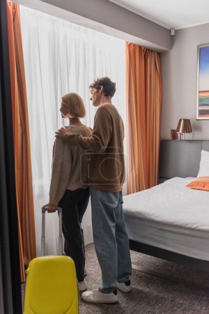 Téléchargez les photos : Young man hugging girlfriend standing with suitcase near window in hotel bedroom - en image libre de droit