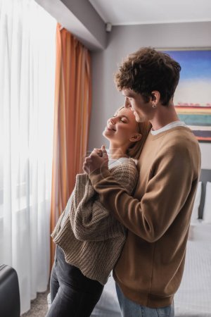 happy blonde woman with closed eyes holding hands with smiling boyfriend in hotel apartments