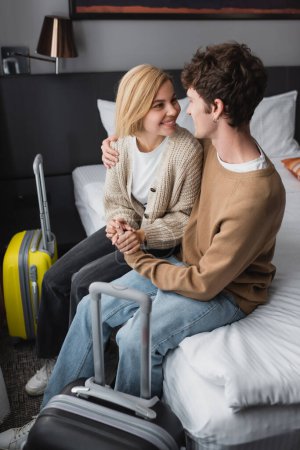 Foto de Cheerful young couple holding hands and looking at each other near travel bags in hotel bedroom - Imagen libre de derechos