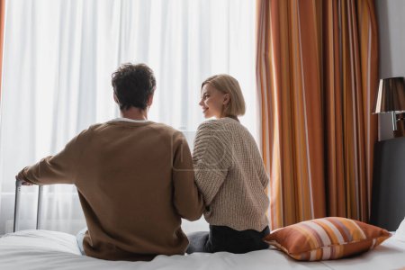 Téléchargez les photos : Back view of happy woman looking at brunette boyfriend while sitting in hotel bedroom - en image libre de droit