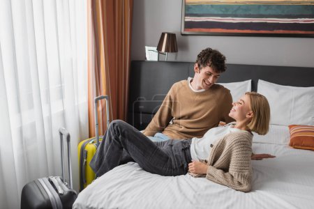 brunette man looking at happy blonde girlfriend on bed in modern hotel bedroom