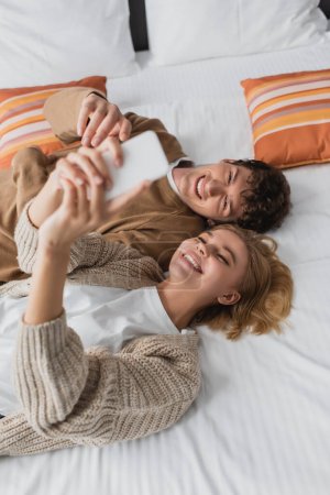 top view of young cheerful couple taking selfie on blurred mobile phone while lying on bed in hotel