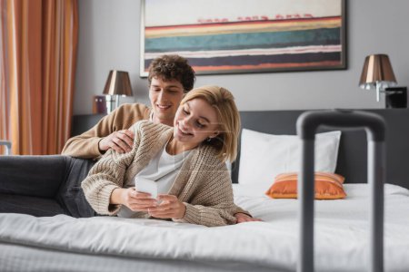 smiling woman using mobile phone near happy young boyfriend on bed in hotel apartments