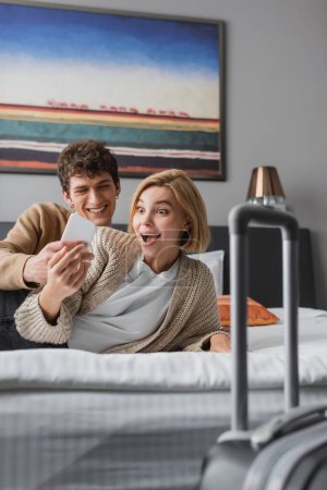 smiling man showing mobile phone to amazed girlfriend on bed in hotel apartments