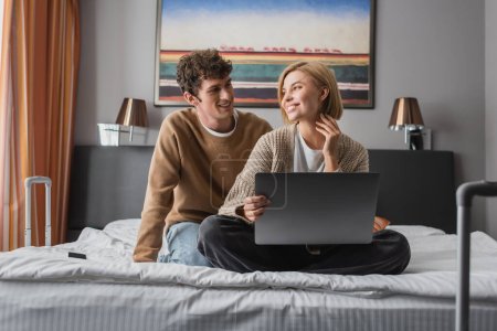 young travelers looking at each other while sitting with laptop in hotel bedroom