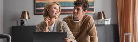 blonde woman and brunette man smiling at each other near laptop in hotel apartments, banner