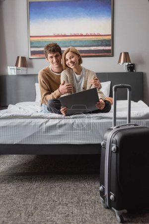 cheerful couple of travelers looking at camera while sitting on bed with laptop near suitcase in hotel room