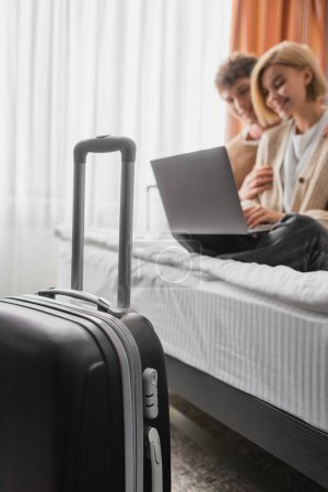 selective focus of travel bag near blurred couple watching movie on laptop in hotel bedroom
