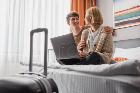 happy young couple looking at each other while sitting with laptop on bed in hotel room 