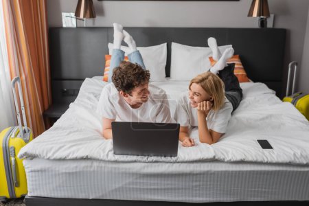 cheerful couple looking at each other while lying on hotel bed near laptop and smartphone with blank screen