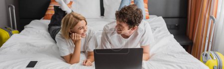 cheerful young couple looking at each other near laptop and smartphone on bed in hotel suite, banner