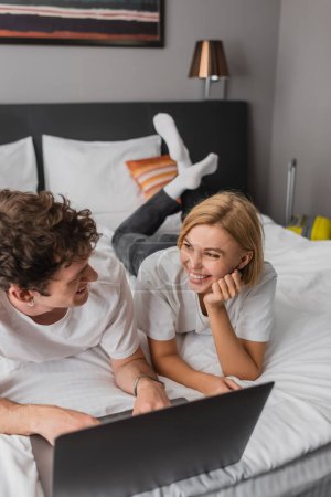 cheerful blonde woman looking at boyfriend near laptop on bed in hotel