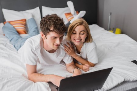 young joyful couple watching film on laptop while lying on bed in hotel apartment