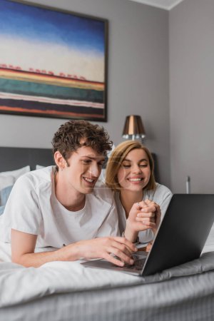 smiling couple holding hands while watching movie on laptop on bed in hotel