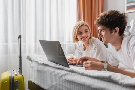 happy blonde woman looking at boyfriend near laptop and travel bag in hotel bedroom