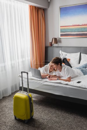 young and happy couple lying on bed and watching film on laptop near suitcase in hotel bedroom