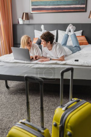young and happy travelers looking at each other on bed near laptop and suitcases on blurred foreground