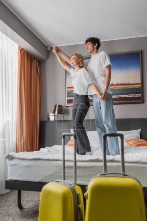 full length of joyful couple standing on bed and holding hands while having fun in hotel room near travel bags