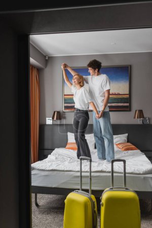 Foto de Couple of young travelers holding hands while having fun on hotel bed near luggage on foreground - Imagen libre de derechos