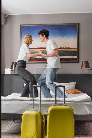 Foto de Side view of excited couple holding hands and having fun on bed near baggage in modern hotel bedroom - Imagen libre de derechos