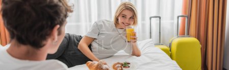 happy woman with glass of orange juice looking at blurred boyfriend on bed in hotel apartments, banner