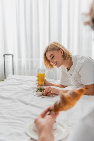 cheerful blonde woman having breakfast with orange juice and croissant on hotel bed near blurred boyfriend