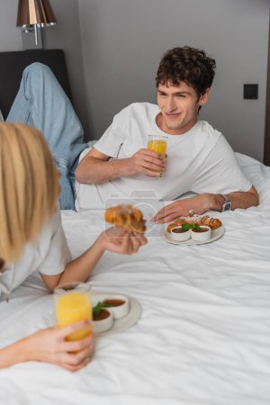 cheerful man holding orange juice and looking at blonde girlfriend having breakfast on hotel bed