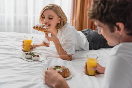 happy woman with orange juice eating tasty croissant near blurred boyfriend on bed in hotel apartments