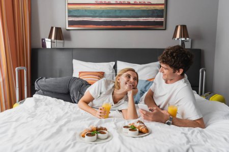 young travelers smiling at each other near orange juice and croissants on hotel bed