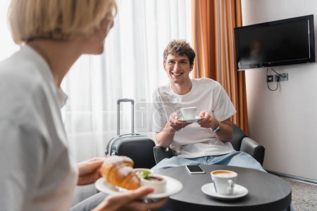 happy man sitting with coffee cup near blurred girlfriend holding croissant while having breakfast in hotel
