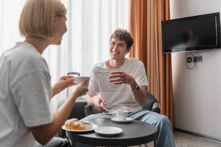 happy man looking at girlfriend drinking morning coffee near croissant in hotel