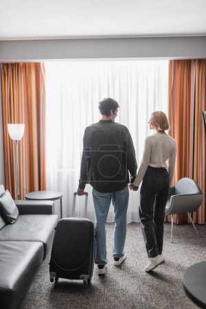 back view of young tourists with suitcase holding hands and looking at each other in hotel room 